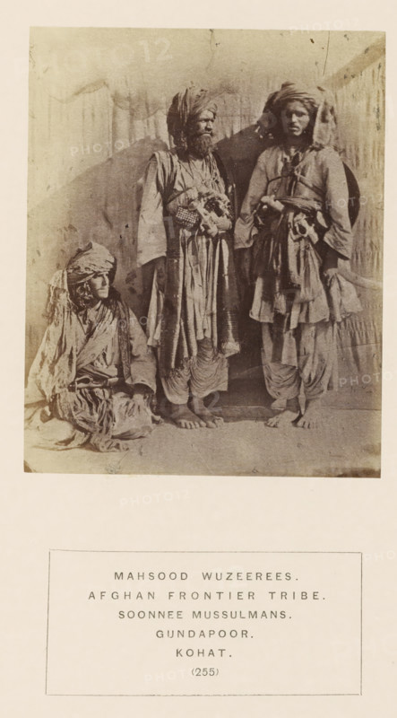 Portrait of three unknown men from the Mahsud tribe of Kohat, Khyber Pakhtunkhwa, Pakistan, photograph, page, inscription: 'MAHSOOD WUZEEREES., AFGHAN FRONTIER TRIBE., SOONNEE MUSSULMANS., GUNDAPOOR., KOHAT.', number: '(255)', Kohat, c. 1862 - in or before 1872, photographer, paper, albumen print, height 111 mm x width 90 mm, head-gear: turban, human races, peoples, nationalities, The sepia photograph shows three individuals wearing layered, loose garments and turbans, with one person seated on the ground to the left and two standing in the center and right; the seated figure has a wrapped head covering and draped cloth around the shoulders, the central standing figure has a full beard, a long outer robe with decorative elements at the waist and layered lower garments, and bare feet visible on the floor, while the right standing figure wears a turban, a belted tunic with hanging fabric and layered lower garments and also has bare feet; a plain backdrop with faint vertical marks and a s...