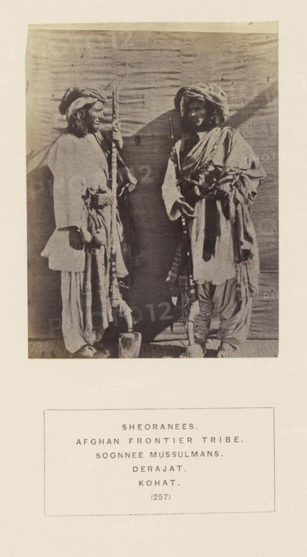 Portrait of two unknown men with masks from the Sherani tribe of Kohat, Khyber Pakhtunkhwa, Pakistan, photograph, page, inscription: 'SHEORANEES., AFGHAN FRONTIER TRIBE., SOONNEE MUSSULMANS., DERAJAT., KOHAT.', number: '(257)', Kohat, c. 1862 - in or before 1872, photographer, paper, albumen print, height 114 mm x width 85 mm, head-gear: turban, firearms: rifle, human races, peoples, nationalities, A sepia-toned photograph shows two standing men facing each other against a plain backdrop. Both wear layered loose garments with draped fabric and turbans on their heads. Each man holds a long, vertical object that reaches near the top of the image. The man on the left stands with his body turned slightly toward the other, one hand resting near his waist and the other gripping his object, and he has visible long hair beneath his turban. The man on the right stands with a relaxed posture, one arm bent and resting across his torso while the other hand holds his object; he has a beard and his ...