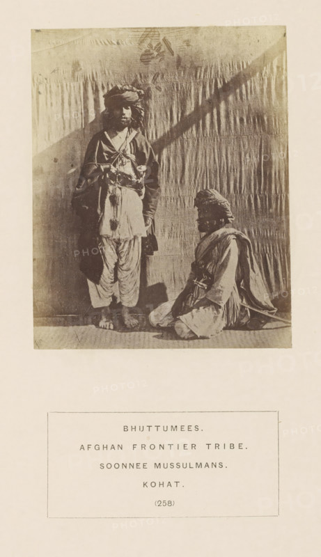 Portrait of two unknown tribesmen with turbans from Kohat, Khyber Pakhtunkhwa, Pakistan, photograph, page, number: '(258)', inscription: 'BHUTTUMEES., AFGHAN FRONTIER TRIBE., SOONNEE MUSSULMANS., KOHAT.', Kohat, c. 1862 - in or before 1872, photographer, paper, albumen print, height 109 mm x width 88 mm, head-gear: turban, human races, peoples, nationalities, A sepia-toned photograph showing two men in traditional clothing against a draped backdrop, one man standing barefoot with layered garments and a turban, the other seated on the floor wearing similar attire and a turban, both facing forward, with the standing man's shadow cast on the backdrop and a rectangular caption box below the photograph.