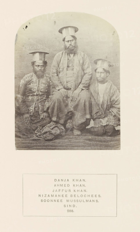 Portrait of Danja Khan, Ahmed Khan and Jaffur Khan, Sindh, Pakistan, photograph, page, Left: Ahmed Kan, center: Danja Khan, right: Jaffur Khan., number: '288.', inscription: 'DANJA KHAN., AHMED KHAN., JAFFUR KHAN., NIZAMANEE BELOCHEES., SOONNEE MUSSULMANS., SIND.', photographer: Henry Charles Baskerville Tanner (possibly), c. 1862 - in or before 1872, paper, albumen print, height 115 mm x width 88 mm, head-gear: hat, ruler, sovereign, human races, peoples, nationalities, A faded sepia photograph shows three seated men wearing tall cylindrical hats with flat tops; the central figure is larger and positioned slightly higher than the other two, with a full beard and hands resting on his knees, wearing layered loose garments and sandals. The man on the left wears a patterned jacket with decorative elements and sits with his hands on his thighs, a folded striped textile at his feet. The man on the right wears a striped outer garment over a lighter inner piece and sits with his hands clasped...