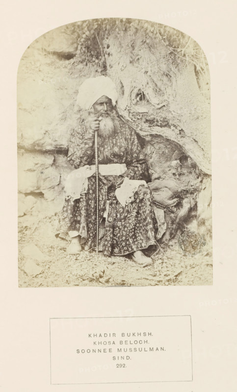 Portrait of Khaddir Bukhsh, a landholder in Sindh, Pakistan, photograph, page, inscription: 'KHADIR BUKHSH., KHOSA BELOCH., SOONNEE MUSSULMAN., SIND.', number: '292.', photographer: Henry Charles Baskerville Tanner (possibly), Sindh, c. 1862 - in or before 1872, paper, albumen print, height 148 mm x width 107 mm, head-gear: turban, human races, peoples, nationalities, A sepia photograph shows an older man seated against a textured rocky background with roots and rough surfaces. He wears a patterned long robe and a large wrapped turban, and he has a long beard and mustache. He holds a long staff with one hand and rests the other hand on his lap, where a light-colored cloth is draped. His shoes are visible beneath the hem of his robe and the ground around him is covered with small stones and debris. The image is framed with rounded top corners and surrounded by a wide light border.