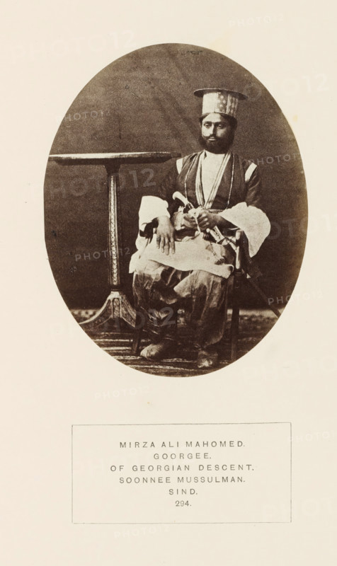 Portrait of Mirza Ali Mahomed, a nobleman from Sindh, Pakistan, photograph, page, number: '294.', inscription: 'MIRZA ALI MAHOMED., GOORGEE., OF GEORGIAN DESCENT., SOONNEE MUSSULMAN., SIND.', photographer: Henry Charles Baskerville Tanner (possibly), Sindh, c. 1862 - in or before 1872, paper, albumen print, height 119 mm x width 94 mm, head-gear: hat, human races, peoples, nationalities, A sepia oval photograph shows a seated man wearing a tall cylindrical hat and layered traditional clothing with wide sleeves and decorative trim, his hands resting in his lap while a sword or curved blade is visible at his side, a small round table with an ornate pedestal stands beside him, and a patterned rug is beneath his feet; the photograph is mounted on a light-colored page with a rectangular label area below the image.