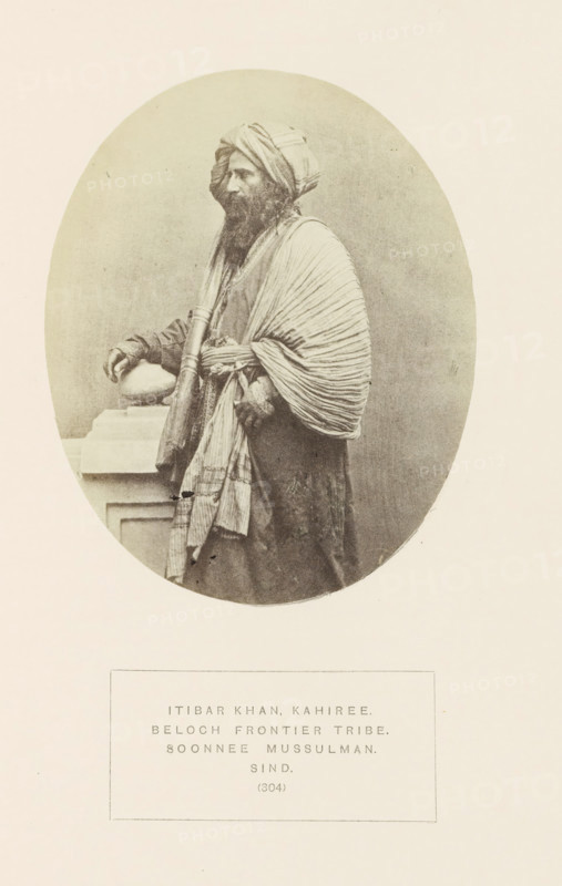Portrait of Itibar Khan, a chief of the Kahiri Baloch tribe in Sindh, Pakistan, photograph, page, inscription: 'ITIBAR KHAR, KAHIREE., BELOCH FRONTIER TRIBE., SOONNEE MUSSULMAN., SIND.', number: '(304)', photographer: Henry Charles Baskerville Tanner (possibly), Sindh, photographer: William Robert Houghton (possibly), c. 1862 - in or before 1872, paper, albumen print, height 128 mm x width 103 mm, ruler, sovereign, head-gear: turban, human races, peoples, nationalities, A sepia-toned oval portrait shows a bearded man in traditional layered garments and a wrapped turban, standing in profile with one hand resting on a stone ledge and the other holding a rolled cloth or staff, his outer draped shawl displaying pronounced folds and pleats and his clothing featuring textured patterns.