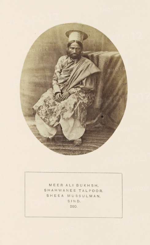 Portrait of Mir Ali Bukhsh, Sindh, Pakistan, photograph, page, inscription: 'MEER ALI BUKHSH., SHAHWANEE TALPOOR., SHEEA MUSSULMAN., SIND, ', inscription: '... Bsu ...', number: '280.', photographer: Henry Charles Baskerville Tanner (possibly), c. 1862 - in or before 1872, paper, albumen print, height 107 mm x width 88 mm, ruler, sovereign, human races, peoples, nationalities, A sepia oval photograph shows a seated person wearing layered garments with patterned fabric and a draped shawl, hands resting together in their lap, a round flat-topped hat on their head, facial hair, and a calm expression, seated beside a draped cloth on a raised surface with visible floorboards beneath.