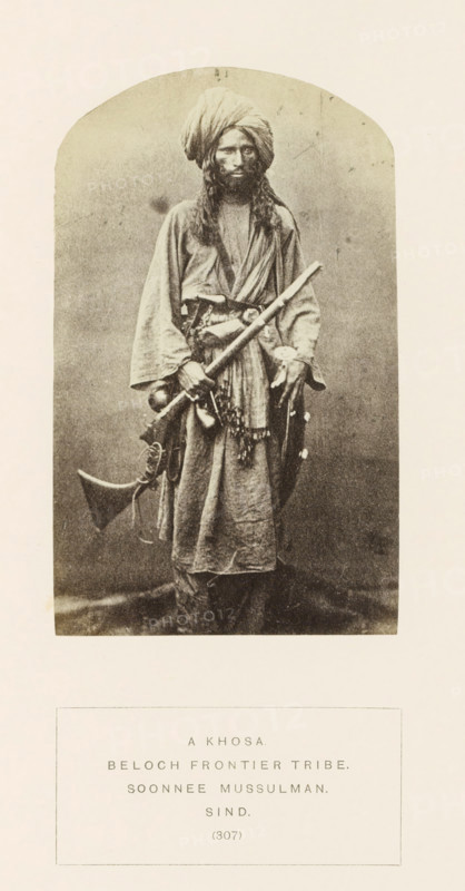 Portrait of an unknown man with a rifle from the Khosa Baloch tribe in Sindh, Pakistan, photograph, page, inscription: 'A KHOSA., BELOCH FRONTIER TRIBE., SOONNEE MUSSULMAN., SIND.', number: '(307)', photographer: Henry Charles Baskerville Tanner (possibly), Sindh, photographer: William Robert Houghton (possibly), c. 1862 - in or before 1872, paper, albumen print, height 129 mm x width 79 mm, head-gear: turban, firearms: rifle, human races, peoples, nationalities, A sepia-toned portrait of a standing man wearing a wrapped turban and long robe, with long hair and a beard, holding a long staff diagonally across his body and carrying a curved rifle and other objects at his waist, set inside an arched vignette on a pale background.