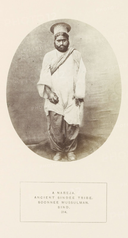Portrait of an unknown man from the Narejo Sindhi tribe in Sindh, Pakistan, photograph, page, inscription: 'A NAREJA., ANCIENT SINDEE TRIBE., SOONNEE MUSSULMAN., SIND.', number: '314.', photographer: Henry Charles Baskerville Tanner (possibly), Sindh, photographer: William Robert Houghton (possibly), c. 1862 - in or before 1872, paper, albumen print, height 129 mm x width 105 mm, head-gear: hat, human races, peoples, nationalities, A sepia-toned oval photograph shows a standing man wearing loose, draped clothing with a long tunic over baggy trousers and flat shoes, a sash or cloth draped diagonally across his chest, and a round cap with a brim; he has a full beard and mustache, dark hair, and a neutral facial expression, with one hand hanging by his side and the other positioned near his thigh, set against a plain backdrop and surrounded by a light border.
