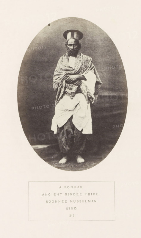 Portrait of an unknown man from the Punar Sindhi tribe in Sindh, Pakistan, photograph, page, inscription: 'A PONHAR., ANCIENT SINDEE TRIBE., SOONNEE MUSSULMAN., SIND.', number: '315.', photographer: Henry Charles Baskerville Tanner (possibly), Sindh, photographer: William Robert Houghton (possibly), c. 1862 - in or before 1872, paper, albumen print, height 139 mm x width 100 mm, head-gear: hat, human races, peoples, nationalities, A sepia-toned oval photograph centered on a pale background shows a standing man facing forward wearing layered traditional clothing and a tall rounded hat; a patterned shawl is draped over his shoulders and crossed arms, a lighter garment hangs around his waist over striped trousers, and he wears light shoes.