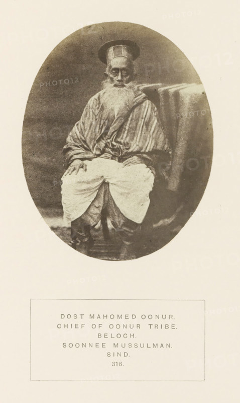 Portrait of Dost Mahomed, a landholder and chieftain from the Baloch community in Sindh, Pakistan, photograph, page, number: '316.', inscription: 'DOST MAHOMED OONUR., CHIEF OF OONUR TRIBE., BELOCH., SOONNEE MUSSULMAN., SIND.', photographer: Henry Charles Baskerville Tanner (possibly), Sindh, photographer: William Robert Houghton (possibly), c. 1862 - in or before 1872, paper, albumen print, height 106 mm x width 86 mm, head-gear: hat, ruler, sovereign, human races, peoples, nationalities, An oval sepia-toned photograph of an elderly man seated facing forward, wearing a tall rounded hat and a long striped robe draped over his shoulders, with a long white beard and hands resting on his knees, light-colored loose trousers gathered over darker footwear, and a textured cloth draped over the back of the chair beside him, all set against a neutral background and mounted on a pale sheet.