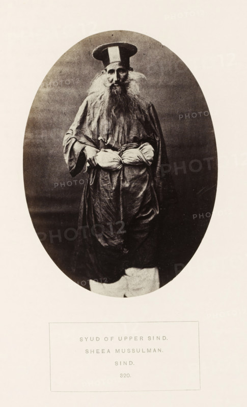 Portrait of an unknown landholder in Sindh, Pakistan, photograph, page, inscription: 'SYUD OF UPPER SIND., SHEEA MUSSULMAN., SIND.', number: '320.', photographer: Henry Charles Baskerville Tanner (possibly), Sindh, photographer: William Robert Houghton (possibly), c. 1862 - in or before 1872, paper, albumen print, height 139 mm x width 101 mm, head-gear: hat, human races, peoples, nationalities, An oval-framed black-and-white photograph shows an elderly man standing and facing forward wearing a tall round hat with a contrasting vertical band, a long flowing robe with wide sleeves, and loose trousers; he has a long full beard and wispy hair at his temples, visible facial features with a solemn expression, and he holds a wrapped bundle at his waist with both hands.
