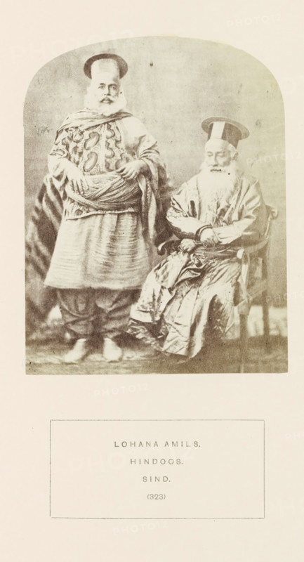 Portrait of two unknown men from the Lohana Amil community in Sindh, Pakistan, photograph, page, number: '(323)', inscription: 'LOHANA AMILS., HINDOOS., SIND.', photographer: Henry Charles Baskerville Tanner (possibly), Sindh, photographer: William Robert Houghton (possibly), c. 1862 - in or before 1872, paper, albumen print, height 123 mm x width 96 mm, head-gear: hat, folk costume, regional costume, human races, peoples, nationalities, A sepia-toned photograph shows two older men wearing traditional garments and tall flat hats; one man stands with hands resting on a wrapped sash around his waist, wearing a patterned tunic and layered skirt-like lower garment with loose trousers beneath, while the other man sits in a wooden armchair with hands folded, wearing a long shiny robe with many folds and a long white beard; the background is plain and the image has rounded top corners and a light border around it.