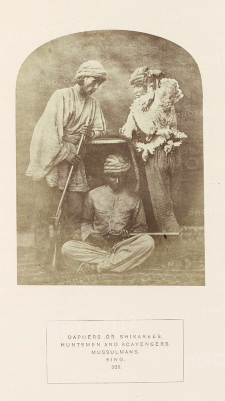 Portrait of three unknown hunters from Sindh, Pakistan, with a dead bird and rifles, photograph, page, number: '326.', inscription: 'DAPHERS OR SHIKAREES., HUNTSMEN AND SCAVENGERS., MUSSULMANS., SIND.', photographer: Henry Charles Baskerville Tanner (possibly), Sindh, photographer: William Robert Houghton (possibly), c. 1862 - in or before 1872, paper, albumen print, height 146 mm x width 108 mm, head-gear: turban, hunter, firearms: rifle, human races, peoples, nationalities, A sepia-toned photograph framed with an arched top shows three men wearing turbans and loose traditional garments; one man stands on the left leaning on a long rifle with both hands, his head tilted downward, another man stands on the right facing slightly toward the center wearing a textured fur or feathered wrap over his shoulders and resting an elbow on a horizontal surface, and a third man sits cross-legged on the ground in front of them holding a long firearm across his lap and looking down; the background is...