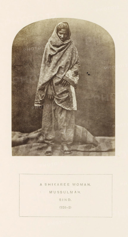 Portrait of an unknown huntress with a headscarf from Sindh, Pakistan, photograph, page, number: '(326-2)', inscription: 'A SHIKAREE WOMAN., MUSSULMAN., SIND.', photographer: Henry Charles Baskerville Tanner (possibly), Sindh, photographer: William Robert Houghton (possibly), c. 1862 - in or before 1872, paper, albumen print, height 116 mm x width 87 mm, head-gear (with NAME), human races, peoples, nationalities, A sepia-toned photograph shows a standing woman draped in a patterned shawl that covers her head and wraps around her body, leaving only part of her face and hands visible; she wears loose trousers and flat footwear, and stands on a textured cloth or backdrop with a plain, slightly mottled background behind her.
