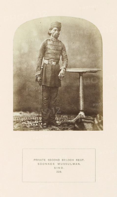 Portrait of a soldier in the second Baloch regiment, Sindh, Pakistan, photograph, page, inscription: 'PRIVATE SECOND BELOCH REGT., SOONNEE MUSSULMAN., SIND.', number: '328.', photographer: Henry Charles Baskerville Tanner (possibly), photographer: William Robert Houghton (possibly), c. 1862 - in or before 1872, paper, albumen print, height 123 mm x width 96 mm, the soldier, the soldier's life, human races, peoples, nationalities, A sepia-toned portrait of a standing man wearing a turban and a long, buttoned tunic with horizontal trim across the chest, a belt with a buckle, straight trousers, and shoes; he rests one hand on a round table with an ornate central pedestal and holds a thin cane in his other hand, standing on a patterned rug against a plain backdrop.
