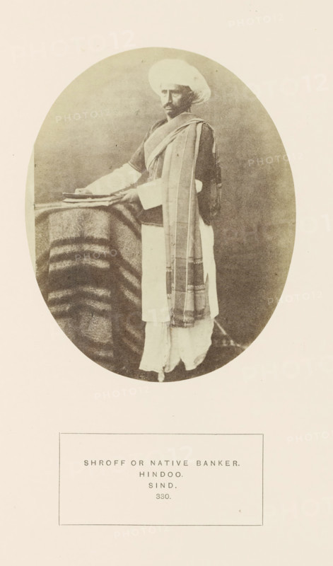 Portrait of an unknown money changer in Sindh, Pakistan, photograph, page, inscription: 'SHROFF OR NATIVE BANKER., HINDOO., SIND.', number: '330.', photographer: Henry Charles Baskerville Tanner (possibly), Sindh, photographer: William Robert Houghton (possibly), c. 1862 - in or before 1872, paper, albumen print, height 129 mm x width 104 mm, head-gear: turban, banker, human races, peoples, nationalities, An oval sepia photograph shows a man standing beside a small table draped with a striped blanket; he wears a wrapped turban and layered garments including a long outer shawl draped over one shoulder and a light-colored lower garment, and his hands rest on a stack of flat objects on the table while his face looks toward the camera.