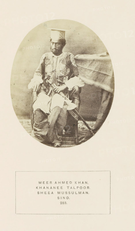 Portrait of Mir Ahmed Khan, Sindh, Pakistan, photograph, page, inscription: 'MEER AHMED KHAN., KHANANEE TALPOOR., SHEEA MUSSULMAN., SIND.', number: '283.', photographer: Henry Charles Baskerville Tanner (possibly), c. 1862 - in or before 1872, paper, albumen print, height 107 mm x width 87 mm, ruler, sovereign, human races, peoples, nationalities, An oval sepia photograph shows a seated man wearing a patterned long coat with decorative embroidery and wide cuffs, striped loose trousers, and a flat square hat; he sits beside a draped table with one hand resting on his knee and the other holding an object, and a sword or long stick leans against the chair extending toward the floor.
