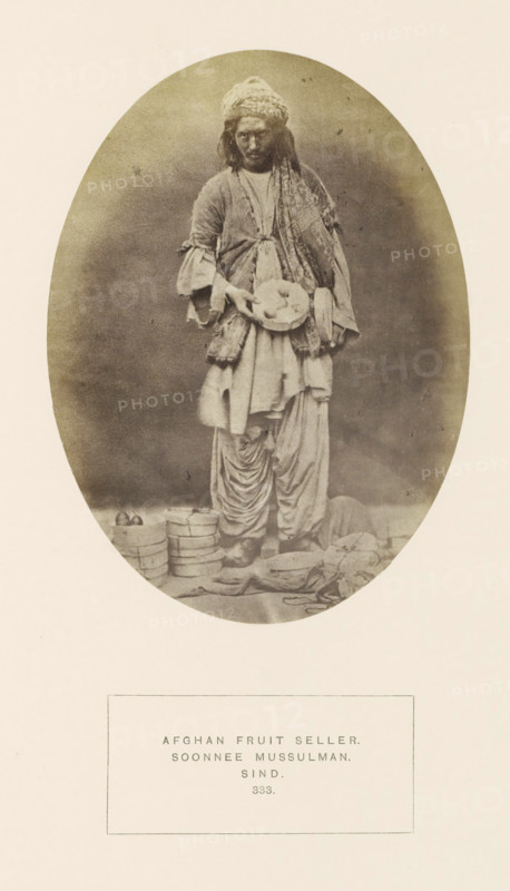 Portrait of an unknown Afghan fruit seller in Sindh, Pakistan, photograph, page, inscription: 'AFGHAN FRUIT SELLER., SOONNEE MUSSULMAN., SIND.', number: '333.', photographer: Henry Charles Baskerville Tanner (possibly), Sindh, photographer: William Robert Houghton (possibly), c. 1862 - in or before 1872, paper, albumen print, height 147 mm x width 108 mm, merchant, salesman, human races, peoples, nationalities, An oval-framed sepia photograph of a standing person wearing layered loose clothing and a patterned headwrap, holding a shallow round tray with round items; the clothing includes a long draped garment, a sash, and pleated lower trousers, with visible worn shoes; at the subject's feet are stacked round containers and scattered cloth bundles and small objects; the background is plain and darker within the oval while the surrounding border is light.