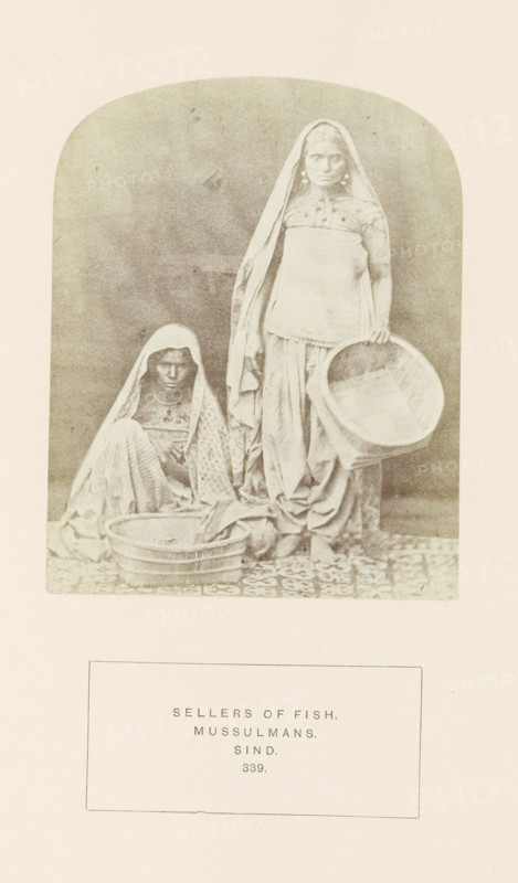 Portrait of two unknown fish sellers in Sindh, Pakistan, photograph, page, inscription: 'SELLERS OF FISH., MUSSULMANS., SIND.', number: '339.', photographer: Henry Charles Baskerville Tanner (possibly), Sindh, photographer: William Robert Houghton (possibly), c. 1862 - in or before 1872, paper, albumen print, height 123 mm x width 97 mm, merchant, salesman, head-gear (with NAME), human races, peoples, nationalities, A faded sepia photograph shows two women in traditional draped garments and head coverings within an arched frame, one woman seated on a patterned floor holding a shallow basket in front of her and the other standing beside her holding a larger round sieve or shallow basket at her side; both wear layered skirts and shawls, and the standing woman has visible earrings.