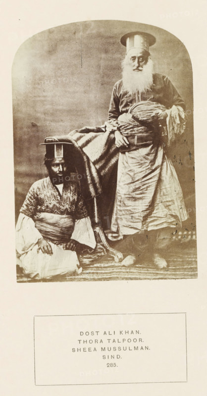 Portrait of Ali Khan and an attendent Sindh, Pakistan, photograph, page, inscription: 'DOST ALI KHAN., THORA TALPOOR., SHEEA MUSSULMAN., SIND.', number: '285.', photographer: Henry Charles Baskerville Tanner (possibly), Sindh, c. 1862 - in or before 1872, paper, albumen print, height 139 mm x width 96 mm, ruler, sovereign, human races, peoples, nationalities, A sepia-toned portrait shows two men in traditional clothing against a plain backdrop, one seated on the floor wearing layered garments with a patterned upper piece and a wrapped sash, a small flat-topped headpiece, and a calm facial expression, the other standing with a long white beard, wearing a long robe and a tall cylindrical hat, holding a wrapped bundle and draped fabric, with a blanket or cloth hanging between them and a patterned rug beneath.