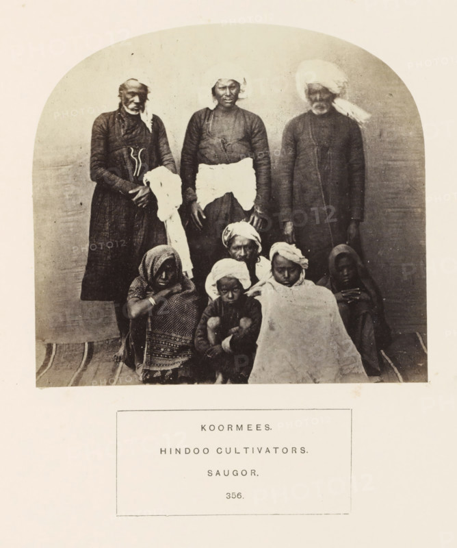 Portrait of eight people and children from the Kurmi community of Sagar, Madhya Pradesh, India, photograph, page, inscription: 'KOORMEES., HINDOO CULTIVATORS., SAUGOR.', number: '356.', photographer: James John Waterhouse (possibly), Sagar, 1862, Central India, paper, albumen print, height 118 mm x width 129 mm, human races, peoples, nationalities, A sepia photograph shows a group portrait of people arranged in two rows against a plain curved backdrop, with three adults standing in the back and several individuals seated or crouched in front; the standing figures wear long dark garments and headwraps, one holds a light cloth, and the central standing person has a contrasting light cloth around the waist, while the seated group features patterned shawls, cloths wrapped around heads, and a large light blanket draped over one person, all on a striped floor covering.