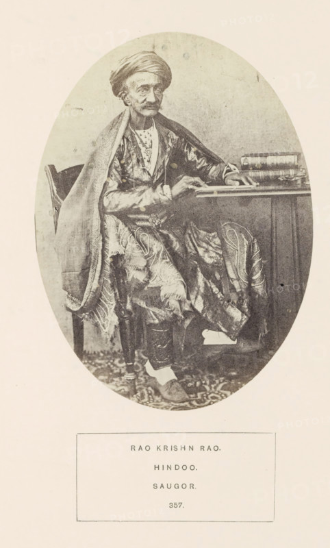Portrait of Rao Krishn Rao, a pundit from Sagar at a reading table, Madhya Pradesh, India, photograph, page, inscription: 'RAO KRISHN RAO., HINDOO., SAUGOR.', number: '357.', photographer: James John Waterhouse (possibly), Sagar, 1862, Central India, paper, albumen print, height 148 mm x width 109 mm, scholar in his study, human races, peoples, nationalities, An oval-framed black-and-white portrait of an older man seated beside a table, wearing a turban and layered patterned robes with a draped shawl over one shoulder, his hands resting on the table and a stack of books beside him, a carved wooden chair partly visible behind him, a patterned floor covering beneath his feet, and a rectangular border containing printed lines below the oval.