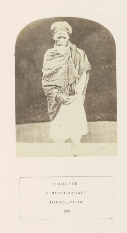 Portrait of Toolsee, a dacoit who was imprisoned in Jabalpur, Madhya Pradesh, India, photograph, page, inscription: 'TOOLSEE., HINDOO DACAIT., JUBBULPOOR.', number: '360.', photographer: James John Waterhouse (possibly), Jabalpur, 1862, Central India, paper, albumen print, height 115 mm x width 84 mm, head-gear: turban, robber, human races, peoples, nationalities, A faded black and white photograph of an elderly person standing inside a rounded rectangular frame, wearing a patterned shawl draped over one shoulder and a wrapped head covering, with hands held together in front and a light-colored garment below the shawl, set against a dark backdrop and pale foreground.
