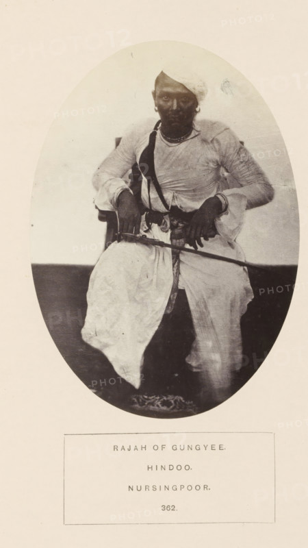 Portrait of an unknown raja in Narsinghpur, Madhya Pradesh, India, photograph, page, number: '362.', inscription: 'RAJAH OF GUNGYEE., HINDOO., NURSINGPOOR.', photographer: James John Waterhouse (possibly), Narsinghpur, 1862, Central India, paper, albumen print, height 139 mm x width 100 mm, ruler, sovereign, head-gear: turban, human races, peoples, nationalities, An oval-framed black-and-white photograph shows a seated person wearing a light-colored turban and layered clothing with long sleeves, a dark strap across the chest, and jewelry at the neck and ears; the person's hands rest on a long thin object held horizontally and the lower garments drape over the knees, with patterned footwear visible at the feet.