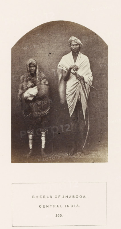 Portrait of a soldier Nar Sing and his wife Chundi from the Bhil tribe of Jhabua, Madhya Pradesh, India, photograph, page, inscription: 'BHEELS OF JHABOOA., CENTRAL INDIA.', number: '365.', photographer: James John Waterhouse (possibly), Jhabua, 1862, Central India, paper, albumen print, height 115 mm x width 79 mm, human races, peoples, nationalities, A sepia photograph framed by an arched top shows a standing man and a seated or slightly crouched person beside him against a plain backdrop; the man is draped in a wrapped cloth over one shoulder and wears a turban, holding a long curved object and another stick-like item across his body, while the person next to him is wrapped in patterned fabric with a hood covering the head and cradling a bundle, and both figures have bare feet with decorative bands visible on the lower legs of the seated person.