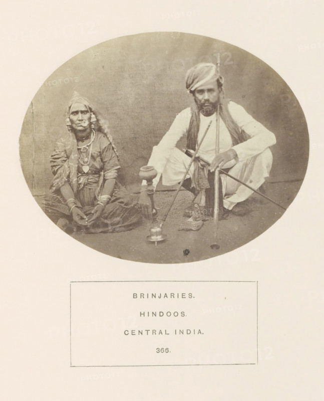 Portrait of two unknown people from the Banjara tribe with a hookah, Madhya Pradesh, India, photograph, page, inscription: 'BRINJARIES., HINDOOS., CENTRAL INDIA.', number: '366.', photographer: James John Waterhouse (possibly), 1862, Central India, paper, albumen print, height 96 mm x width 120 mm, water-pipe, hookah, narghile, human races, peoples, nationalities, A circular sepia photograph shows a seated man and woman on a plain floor against a neutral backdrop; the woman on the left wears layered traditional garments and headscarf, jewelry including large earrings and necklaces, and sits cross-legged with hands resting in her lap, while the man on the right wears light-colored clothing and a turban, squats with one arm resting on his knee and holds a long staff or pipe that connects to a tall vessel placed between them.