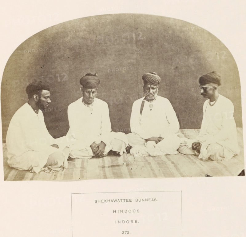 Portrait of four unknown merchants originally from Shekhawati, Rajasthan, based in Indore, Madhya Pradesh, India, photograph, page, number: '372.', inscription: 'SHEKHAWATTEE BUNNEAS., HINDOOS., INDORE.', photographer: James John Waterhouse (possibly), Indore, 1862, Central India, paper, albumen print, height 115 mm x width 168 mm, head-gear: turban, human races, peoples, nationalities, A sepia photograph shows four men seated on a striped cloth floor against a plain curved backdrop, all wearing light-colored tunics and headwraps, with two men facing inward toward the center and the two outer men turned slightly inward; their hands rest on their laps or knees and one man wears a pendant at his chest, while a folded cloth lies in front of them and the image has a worn, aged appearance.