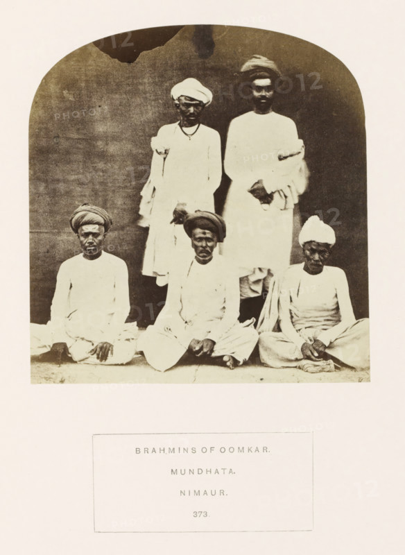 Portrait of five unknown priests (brahmins) from Mandhata, Madhya Pradesh, India, photograph, page, Oomkar from the original title refers to the Omkareshwar Temple located in Mandhata., inscription: 'BRAHMINS OF OOMKAR., MUNDHATA., NIMAUR.', number: '373.', photographer: James John Waterhouse (possibly), Mandhata, 1862, Central India, paper, albumen print, height 125 mm x width 119 mm, head-gear: turban, religious functionaries, Hinduism, Buddhism, Jainism, human races, peoples, nationalities, A sepia-toned photograph framed by an arched background shows five seated and standing men wearing light-colored traditional garments and turbans; two men stand behind three men who sit cross-legged on the ground, all facing forward with neutral expressions, hands resting in laps or clasped, and a plain dark backdrop behind them with a lighter border surrounding the image.