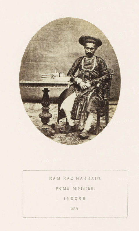 Portrait of Ram Rao Narrain, a prime minister of the princely state of Holkar, Indore, Madhya Pradesh, India, photograph, page, inscription: 'RAM RAO NARRAIN., PRIME MINISTER., INDORE.', number: '388.', photographer: James John Waterhouse (possibly), 1862, Central India, paper, albumen print, height 93 mm x width 75 mm, prime minister, human races, peoples, nationalities, An oval sepia photograph shows a seated man wearing a rounded turban and layered clothing, including a patterned shawl draped over one shoulder and decorative necklaces; he sits on a carved wooden chair with one hand resting on his lap and the other near a small table beside him, which holds stacked books and a small container; the table has an ornate pedestal base and the scene is set against a plain backdrop with a patterned floor covering visible beneath the chair and table.