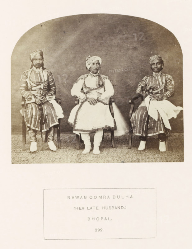 Portrait of Oomra Dulha, husband of Sekunder Begum, a queen of Bhopal, Madhya Pradesh, India, photograph, page, inscription: 'NAWAB OOMRA DULHA., (HER LATE HUSBAND.), BHOPAL.', number: '392.', photographer: James John Waterhouse (possibly), Bhopal, 1862, Central India, paper, albumen print, height 102 mm x width 118 mm, ruler, sovereign, human races, peoples, nationalities, A sepia-toned photograph shows three seated men wearing patterned robes and turbans, each sitting on a wooden chair against a plain backdrop; the central figure wears a lighter outfit with decorative trim and has hands folded in his lap, while the two flanking figures wear striped garments and hold canes or staffs with cloth draped over their arms, all three wearing footwear and positioned on a patterned floor covering.