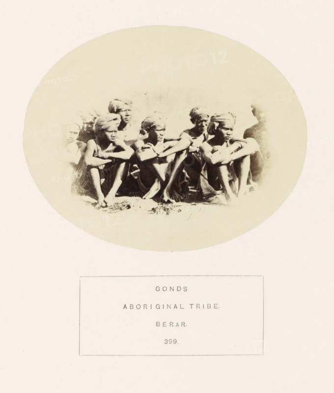Group portrait of people from the Gond tribe of the former Berar in central India, photograph, page, number: '399.', inscription: 'GONDS., ABORIGINAL TRIBE., BERAR.', photographer: James Mulheran (possibly), Berar, 1862, paper, albumen print, height 95 mm x width 120 mm, human races, peoples, nationalities, A faded oval photograph shows a group of people sitting closely together with arms resting on their knees; they wear headwraps and simple clothing, their skin tones rendered in sepia, and the image has a soft, high-contrast look with faces and limbs partially obscured by light areas and shadow.