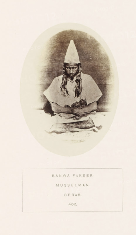 Portrait of an unknown fakir from the former Berar in central India, photograph, page, number: '402.', inscription: 'BANWA FAKEER., MUSSULMAN., BERAR.', photographer: James Mulheran (possibly), Berar, 1862, paper, albumen print, height 103 mm x width 77 mm, human races, peoples, nationalities, asceticism, Hinduism, Buddhism, Jainism, An oval sepia photograph shows a person seated facing forward wearing a tall conical hat and a loose, light-colored garment with short sleeves; the person's long hair falls over the shoulders and they look downward with hands resting in their lap holding a cloth or bundle, and a rectangular object lies on the surface in front of them, all set against a softly shaded background within the oval, surrounded by a pale border and further pale margins.
