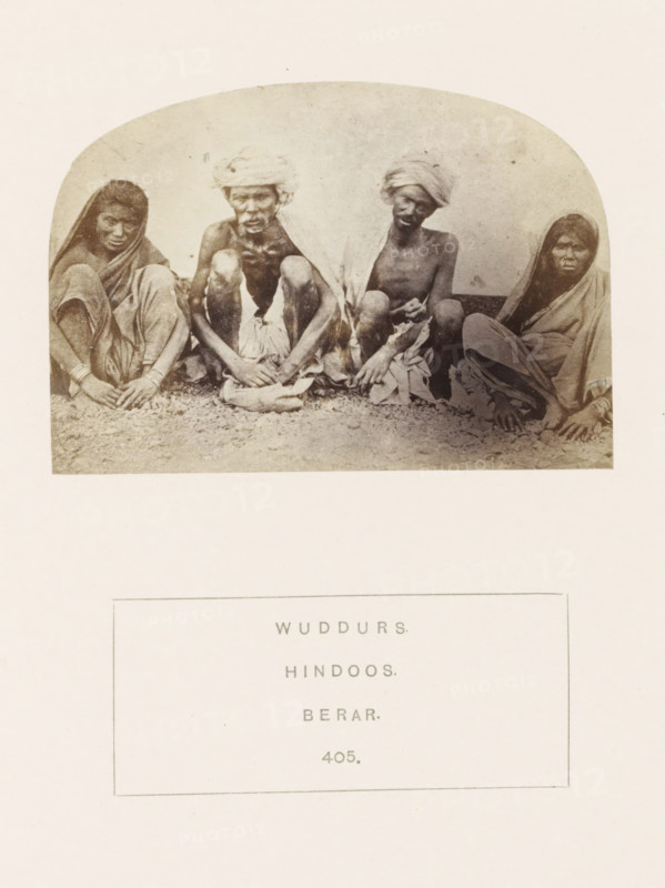 Portrait of four unknown people from the Waddar tribe of the former Berar in central India, photograph, page, inscription: 'WUDDURS., HINDOOS., BERAR.', number: '405.', photographer: James Mulheran (possibly), Berar, 1862, paper, albumen print, height 66 mm x width 95 mm, human races, peoples, nationalities, A faded oval photograph shows four seated people on a textured ground against a plain background, two men in the center wearing cloths wrapped around their heads and shoulders and two women at the sides draped in shawls, all sitting with legs drawn close and hands resting near their feet, wearing simple garments and appearing in sepia tones with visible grain and slight discoloration around the edges.