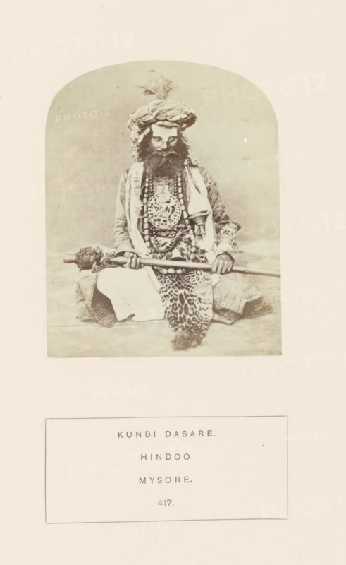 Portrait of an unknown man from the Kunbi community with ceremonial clothes, Mysore, Karnataka, India, photograph, page, inscription: 'KUNBI DASARE., HINDOO., MYSORE.', number: '417.', c. 1865 - in or before 1875, photographer, South India, paper, albumen print, height 95 mm x width 75 mm, head-gear: turban, human races, peoples, nationalities, ritual practices with particular objectives, Hinduism, Buddhism, Jainism, A sepia-toned photograph framed by a rounded rectangular border shows a seated man wearing a large wrapped turban with a plume, a full beard, and layered garments including a patterned robe and a draped cloth; he holds a long staff horizontally across his lap with both hands, and several beaded necklaces and a large ornate medallion hang over his chest, while a patterned textile is draped over his legs and he sits on a flat surface against a plain backdrop.
