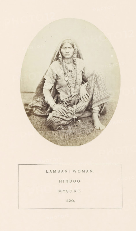 Portrait of an unknown woman from the Lambani tribe of Mysore, Karnataka, India, photograph, page, The Lambani tribe is also known as Banjara., number: '420.', inscription: 'LAMBANI WOMAN., HINDOO., MYSORE.', Mysore, c. 1865 - in or before 1875, photographer, South India, paper, albumen print, height 98 mm x width 76 mm, head-gear (with NAME), human races, peoples, nationalities, A sepia-toned oval photograph shows a seated woman wearing a head covering and layered necklaces, with patterned trousers and a sash tied at the waist; she sits barefoot on a textured mat with one knee raised and hands resting on her legs, her expression neutral and her hair partially visible beneath the veil, and the photograph is set on a pale background with a rectangular bordered area below.
