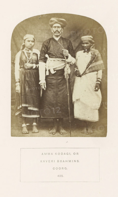 Portrait of three people from the Kodava community of Kodagu. Karnataka, India, photograph, page, number: '425.', inscription: 'AMMA KODAGI, OR, KAVERI BRAHMINS., COORG.', photographer: G. Richter (possibly), Kodagu, c. 1865 - in or before 1875, South India, Karnataka, paper, albumen print, height 122 mm x width 95 mm, human races, peoples, nationalities, A sepia photograph shows three barefoot people standing side by side against a plain backdrop, with a man in the center wearing a dark long garment, a wrapped sash at his waist, a turban, and holding a long staff while a curved knife rests at his side; a girl on his left wears a dress with decorative trim, multiple necklaces, bracelets, and anklets, and has a cloth tied around her head; a person on his right wears a light wrapped garment with a shawl draped over one shoulder and a head covering, and also wears bracelets.
