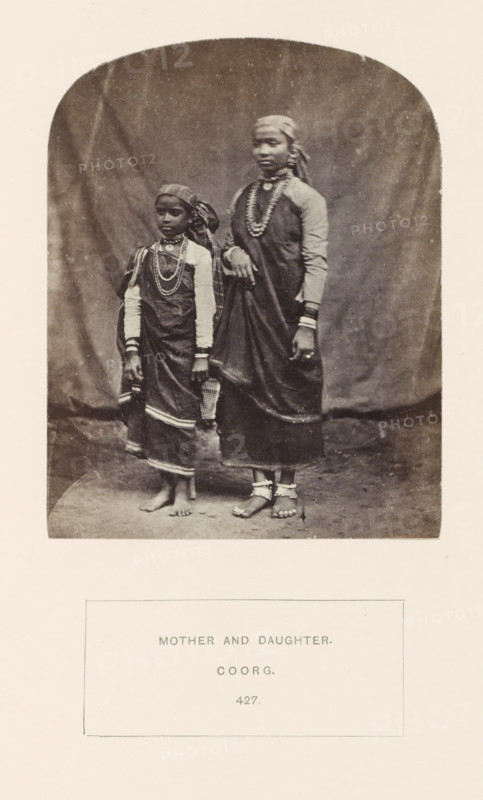 Portrait of an unknown mother and daughter from the Kodava community of Kodagu, Karnataka, India, photograph, page, number: '427.', inscription: 'MOTHER AND DAUGHTER., COORG.', photographer: G. Richter (possibly), Kodagu, c. 1865 - in or before 1875, South India, paper, albumen print, height 123 mm x width 97 mm, mother and daughter(s) (family group), human races, peoples, nationalities, A sepia-toned photograph shows two females standing on a plain floor against a draped fabric backdrop; both wear long dark garments with lighter long-sleeved underlayers and patterned headscarves, and both are adorned with multiple layered necklaces and bracelets; the taller figure rests one hand on the shorter figure’s shoulder and wears anklets and toe rings while the shorter figure’s bare feet are visible and she has a cloth draped over one arm; the image has rounded top corners and is mounted on a lighter paper.