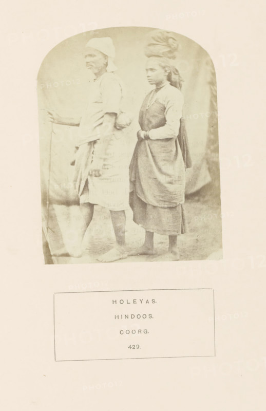 Portrait of an unknown man and woman from the Holeya community of Kodagu, Karnataka, India, photograph, page, inscription: 'HOLEYAS., HINDOOS., COORG.', number: '429.', photographer: G. Richter (possibly), Kodagu, c. 1865 - in or before 1875, South India, paper, albumen print, height 115 mm x width 88 mm, human races, peoples, nationalities, A faded sepia photograph shows two barefoot individuals standing side by side against a plain backdrop, the person on the left facing sideways and holding a long, thin staff while wearing a short-sleeved garment with a wrapped lower cloth and a head covering, and the person on the right standing slightly behind with hands clasped in front, wearing layered garments including a sleeveless outer piece over a long-sleeved top and a wrapped lower skirt, with a cloth wrapped around their head and hair visible at the back.