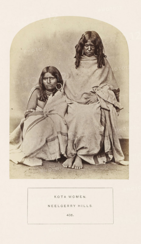 Portrait of two unknown women from the Kota tribe of the Nilgiri mountains in Tamil Nadu, India, photograph, page, inscription: 'KOTA WOMEN., NEELGERRY HILLS.', number: '436.', Nilgiris, c. 1865 - in or before 1875, photographer, South India, Tamil Nadu, paper, albumen print, height 145 mm x width 107 mm, human races, peoples, nationalities, A sepia-toned photograph shows two seated women wrapped in plain draped cloths, one sitting slightly lower and leaning against the other, both with long dark hair and large circular earrings; the taller woman has her hands folded over her lap and bare feet visible at the bottom edge of the drapery, while the shorter woman wears multiple necklaces and bracelets and looks slightly away from the camera; the background is a plain neutral backdrop and the image is bordered by a rounded top frame.