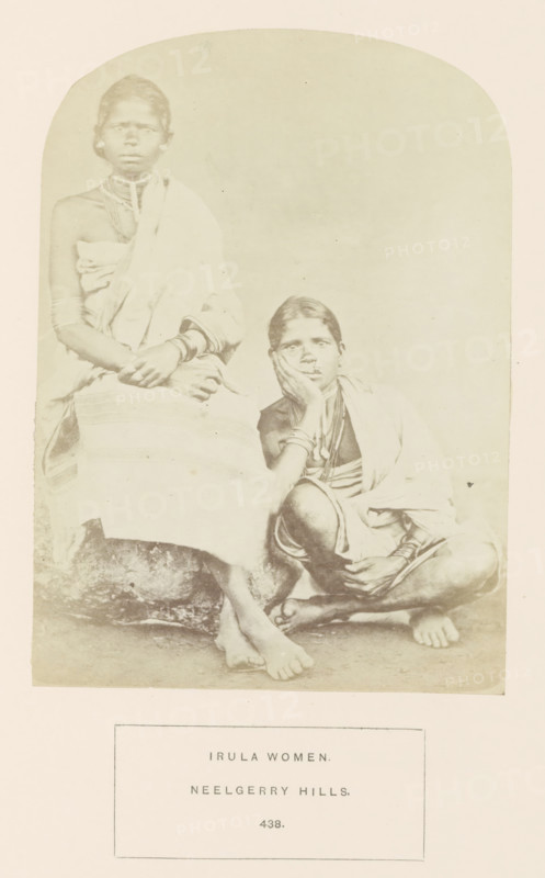 Portrait of two unknown women from the Irula tribe of the Nilgiri mountains in Tamil Nadu, India, photograph, page, number: '438.', inscription: 'IRULA WOMEN., NEELGERRY HILLS.', Nilgiris, c. 1865 - in or before 1875, photographer, South India, Tamil Nadu, paper, albumen print, height 165 mm x width 119 mm, human races, peoples, nationalities, A faded sepia photograph shows two seated women wearing draped cloth garments and multiple necklaces and bracelets; one woman sits upright with her hands resting in her lap while the other sits lower with an elbow propped on a knee and her hand supporting her face, both barefoot on a textured ground, with a plain light background and a rectangular bordered panel beneath the image.