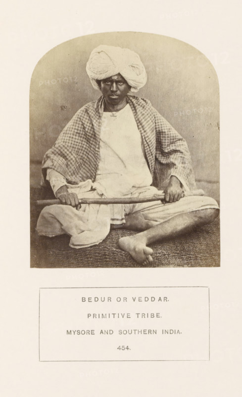 Portrait of an unknown man with a stick in his hands, Mysore, Karnataka, India, photograph, page, number: '454.', inscription: 'BEDUR OR VEDDAR., PRIMITIVE TRIBE., MYSORE AND SOUTHERN INDIA.', c. 1865 - in or before 1875, photographer, South India, paper, albumen print, height 107 mm x width 86 mm, head-gear: turban, human races, peoples, nationalities, A seated person wearing a wrapped turban and a light garment with a checked shawl draped over the shoulders is shown in a framed oval photograph; the person holds a long stick across their lap with both hands and sits cross-legged on a textured mat, with one bare foot visible, and a neutral backdrop behind them.