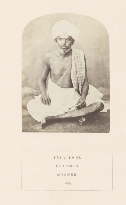 Portrait of an unknown priest from the Vaishnav Brahmin community of Mysore, Karnataka, India, photograph, page, number: '410.', inscription: 'SRI VISHNU., BRAHMIN., MYSORE.', Mysore, c. 1865 - in or before 1875, photographer, South India, paper, albumen print, height 102 mm x width 76 mm, Brahma (supreme being, creator, etc., Hinduism, Buddhism, Jainism), human races, peoples, nationalities, A sepia-toned photograph of an older man seated cross-legged on a plain surface, wearing a light-colored wrapped cloth around his lower body and a turban on his head, with a patterned cloth draped over one shoulder; he is bare-chested and has a thin beard and mustache, with his hands resting on his knees and one hand slightly raised, looking forward, and the image is framed by a rounded-arch border on a light background.