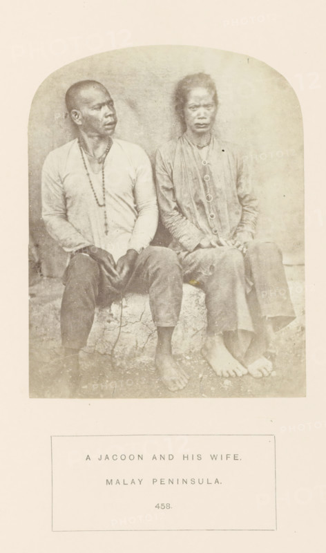 Portrait of an unknown man and woman from the Jakun tribe of Malacca, Malaysia, photograph, page, number: '458.', inscription: 'A JACOON AND HIS WIFE., MALAY PENINSULA.', Malacca, c. 1865 - in or before 1875, photographer, paper, albumen print, height 123 mm x width 97 mm, human races, peoples, nationalities, A sepia-toned photograph shows two seated adults on a low stone or concrete block against a plain background, both barefoot and facing forward; the person on the left wears a light long-sleeved shirt and dark trousers with a beaded necklace hanging down and has a closely shaved head, while the person on the right wears a darker long-sleeved garment with visible round buttons down the front, has longer hair tied back, and holds hands together on their lap.