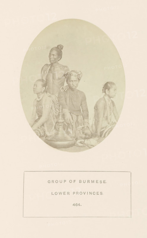 Group portrait of people from Myanmar, photograph, page, number: '464.', Myanmar, c. 1865 - in or before 1875, photographer, paper, albumen print, height 96 mm x width 77 mm, human races, peoples, nationalities, An oval sepia photograph showing four seated and standing figures with darker skin tones arranged closely together, two younger individuals sitting on either side and two adults in the center, one adult seated wearing a patterned headwrap and long-sleeved top and the other adult standing bare-chested with a cloth draped over one shoulder and hair tied in a topknot; the seated figures wear wrapped lower garments and jewelry around their necks, and there is a large lidded ceremonial-looking vessel placed on the floor in front of them.