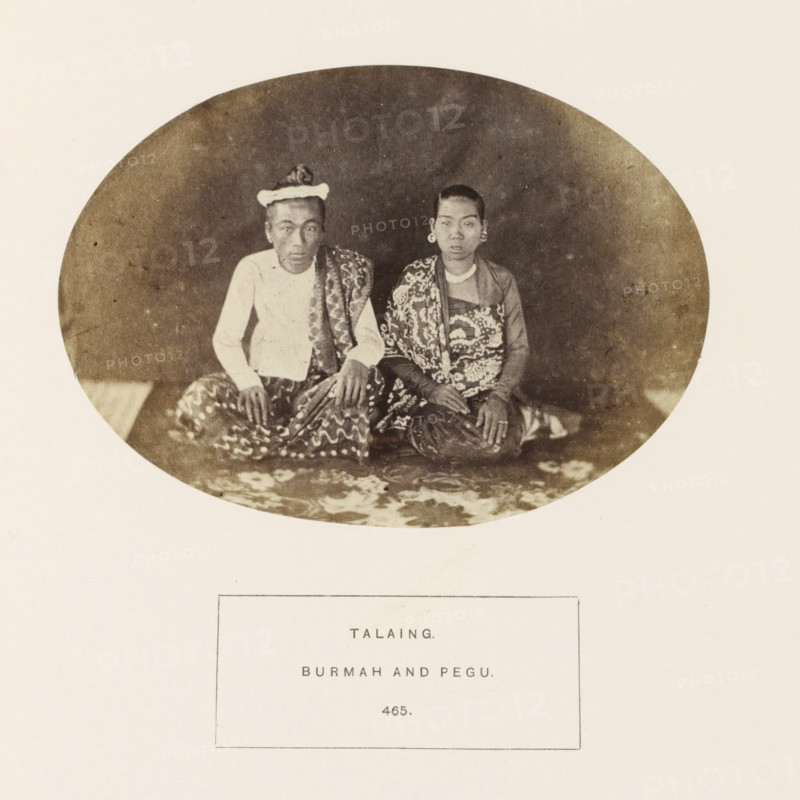 Portrait of a man and woman from the Mon tribe of Myanmar, photograph, page, number: '465.', inscription: 'TALAING., BURMAN AND PEGU.', Myanmar, c. 1865 - in or before 1875, photographer, paper, albumen print, height 98 mm x width 138 mm, human races, peoples, nationalities, An oval sepia photograph shows two seated individuals on a patterned floor covering against a dark backdrop; the person on the left wears a light-colored long-sleeved top, a patterned sarong, and a headwrap, with hands resting on their knees, while the person on the right has short hair, large circular ear ornaments, a patterned garment draped over one shoulder, and hands resting together on their lap.