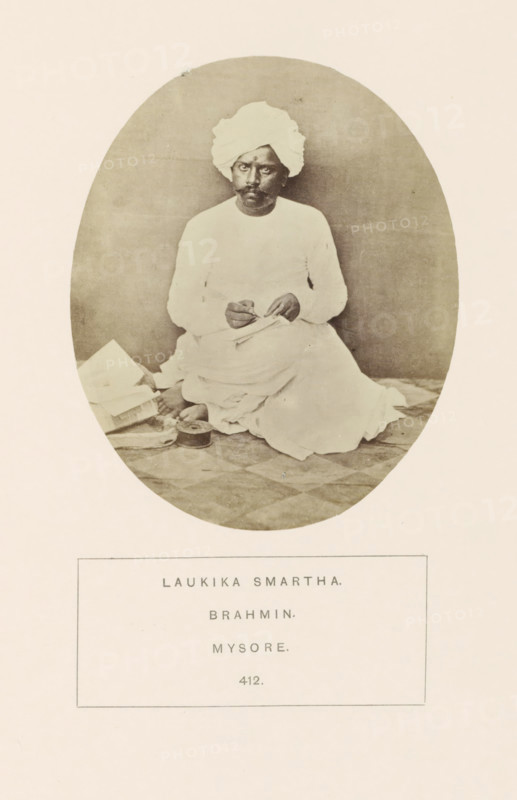 Portrait of an unknown priest from the Smarta Brahmin community of Mysore, Karnataka, India, photograph, page, number: '412.', inscription: 'LAUKIKA SMARTHA., BRAHMIN., MYSORE.', Mysore, c. 1865 - in or before 1875, photographer, South India, paper, albumen print, height 101 mm x width 87 mm, Brahma (supreme being, creator, etc., Hinduism, Buddhism, Jainism), human races, peoples, nationalities, An oval-framed sepia photograph of a man seated on a patterned floor, wearing a light-colored turban and matching long-sleeved garment with draped cloth around his legs; he holds a small object between his hands and gazes forward with a neutral expression; beside him on the floor are several folded papers or books and a round container, and the background is plain and softly shaded.