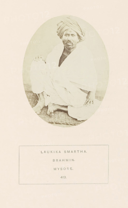 Portrait of an unknown priest from the Smarta Brahmin community of Mysore, Karnataka, India, photograph, page, number: '413.', inscription: 'LAUKIKA SMARTHA., BRAHMIN., MYSORE.', Mysore, c. 1865 - in or before 1875, photographer, South India, paper, albumen print, height 98 mm x width 76 mm, head-gear: turban, Brahma (supreme being, creator, etc., Hinduism, Buddhism, Jainism), human races, peoples, nationalities, An oval-framed sepia photograph shows a seated man wearing a wrapped turban and draped cloth garments, sitting cross-legged on a textured mat with one hand resting on his knee and the other on the mat; his face has a beard and he looks toward the camera, and the image is centered on a pale background with a rectangular bordered area below the oval.