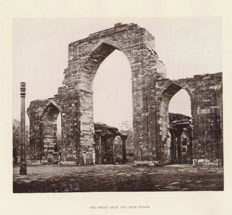 View of a gate and pillar at the Qutb complex in Delhi, The great arch and iron pillar, page, photomechanical print, photographer: Bourne & Shepherd, Delhi, c. 1860 - c. 1877, printmaker, paper, Woodburytype, height 146 mm x width 177 mm, arch, archivolt, architecture, column, pillar, A sepia-toned photograph of a large ornate stone archway and adjacent ruins, showing detailed carved patterns along the arch and supporting pillars, with visible masonry blocks and decorative motifs on vertical surfaces; a tall slender column stands to the left of the arch, and smaller arches and stone structures are visible through and beside the main arch; two figures in light clothing stand beneath the arch near its base; the ground in front is paved with irregular stone slabs and scattered stones, and trees and vegetation are visible through openings among the ruins.